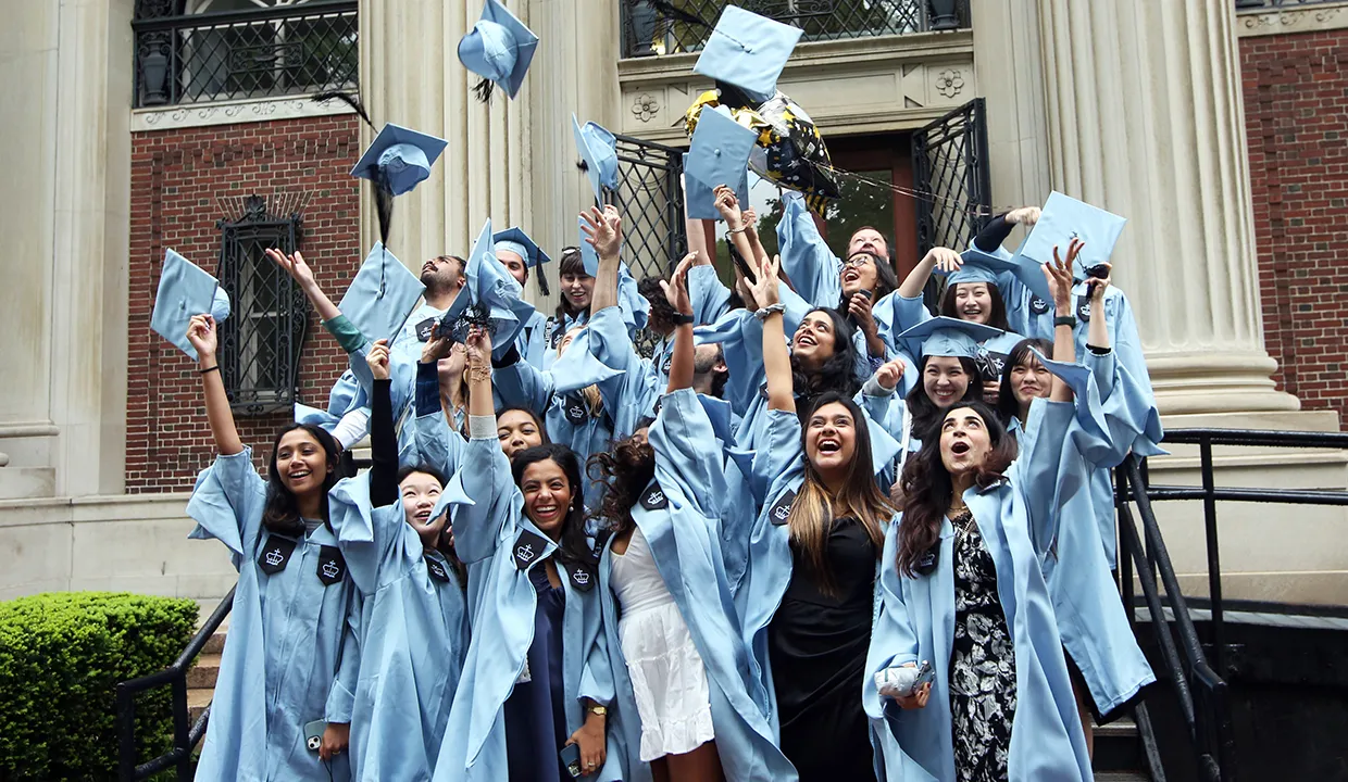 Columbia students throw mortar boards in the air in front of Avery Hall.